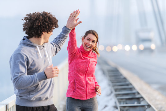 Happy Caucasian runners giving high five during running on the bridge on cold weather. Snow all around, wintertime.