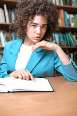 Young beautiful curly girl in glasses and blue suit sitting with books in the library. Student Study