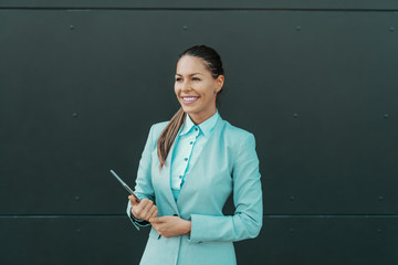 Smiling gorgeous Caucasian woman in formal wear holding tablet and posing in front of dark background outdoors.