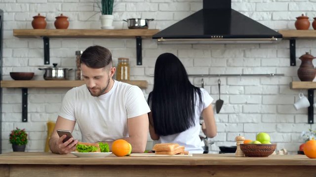 Young couple at the kitchen.  Girlfriend doing morning coffee and giving cup to the guy. He holding smartphone and looking at it.