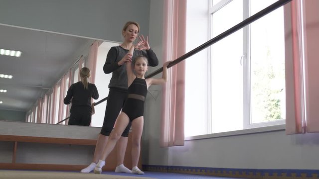 Little gymnast performing gymnastic exercises at wall mounted ballet barre under the instructor control (Grand battement to the side).