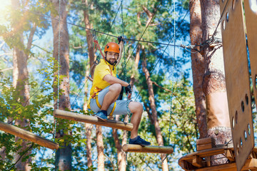 Young smiling man, in protective gear, is sitting on rope trail, bridge on high trees in forest. Rope adventure park with difficult obstacles and ziplines. Extreme rest and summer activities concept.