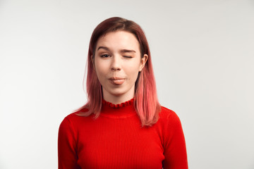 Fototapeta premium A funny girl in glasses makes faces, sticking her tongue out and winks. The concept of frivolous fun and teasing. Isolated on white background in studio