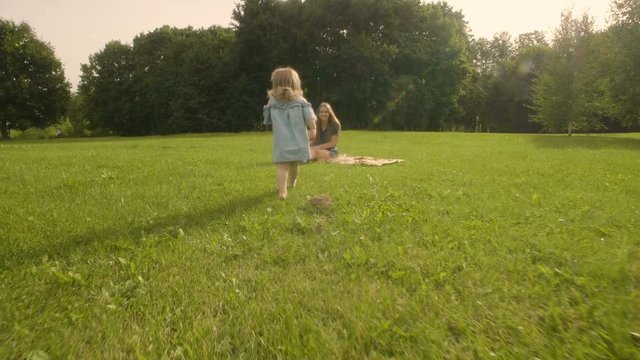 Baby girl running through the grass and rushes into mother hands in garden in sunset sunlight. Slow motion. Happy smiling baby girl.
