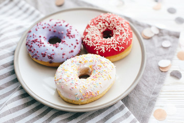 Delicious colorful donuts with sprinkles on stylish plate on white table with confetti. Party and celebration concept. No diet. Candy bar at wedding reception. Purple and pink donuts