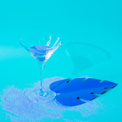 Cocktail glass with tropical leaf in blue colors. Strong fashion shadows. Concept of luxury relax with beverage on the beach.