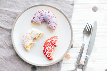 Delicious bite of colorful donuts with sprinkles on stylish plate on white table with confetti, fork and knife, flat lay. Party concept. No diet. Candy bar at wedding reception.