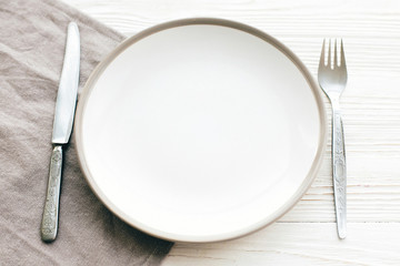 Stylish empty plate with vintage fork and knife on napkin on white table, top view. Modern set, serving for reception and celebration. Party and diet concept
