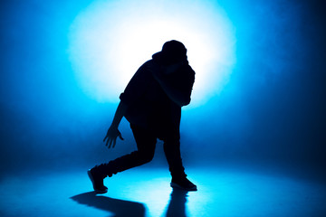 Stylish young man wearing trendy streetwear is dancing contemporary street dance on a blue studio background