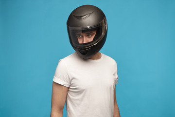 Portrait of stylish young man wearing white t-shirt and motorcycle helmet posing isolated at blue studio wall looking away. Motor sports, extreme lifestyle, transportation and people concept - obrazy, fototapety, plakaty