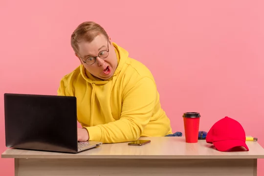 Cute comic man in bright colors casual wear geek working on computer with funny stupid facial expression