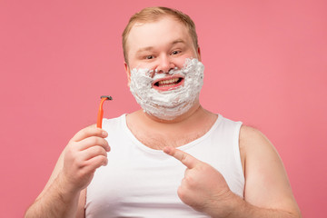 Studio portrait of funny tubby middle-aged man in undershirt smiling at camera with happy joyful expression while shaving his face with razor and foam