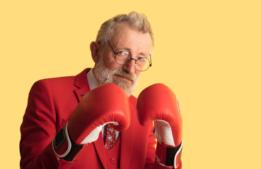 Mature business man in red formal wear wearing boxing gloves posing isolated on yellow background looking at camera with confident expression