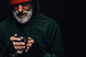 Old homeless man with grey beard covering up in green decrepit wear holding a mug of hot tea to...