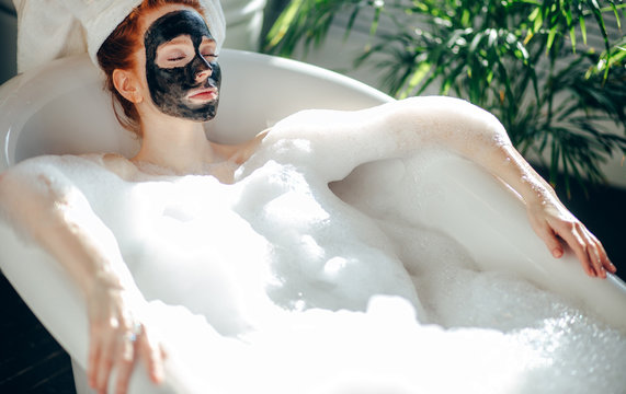 Skin care - Young lady with facial black clay mask applyed on her face relaxing in foam bath, getting rejuvenation spa and beaty procedure at home