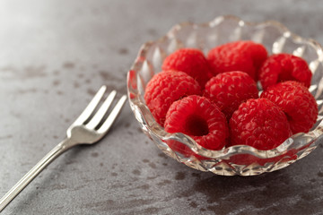 Freshly picked raspberries in a small glass bowl with a fruit fork to the side on a gray background