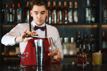 Barman making alcoholic drink, pouring liquid from brassy jigger into the shaker on the bar barstand