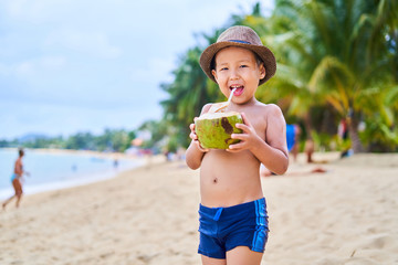 Tanned Asian boy stands on the beach in a hat and drinks coconut