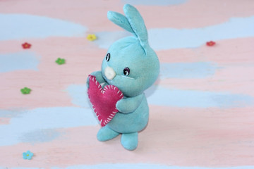 Blue cute bunny with a red heart in its paws on a pink- light blue background. Handwork.