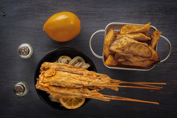 baked fish on skewers in a black plate, french fries and lemon on a wooden table. Close-up. Top view