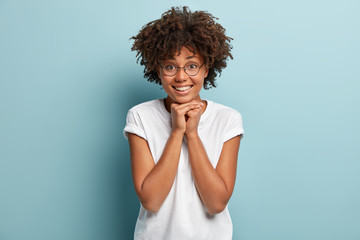 Headshot of satisfied black lady with Afro hairstyle, keeps both hands under chin, smiles cheerfully, hears positive story, dressed in casual clothes, isolated over blue background. Happiness