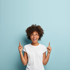 Photo of cheerful lovely woman has Afro haircut, points above at free space, gets good impression of promotion, wears white t shirt and spectacles, isolated over blue background. Look upwards