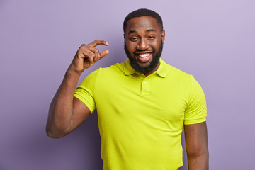 Cheerful black man with bright smile, shows white teeth, shapes something very tiny with hand,...