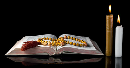 Opened Holy Bible with rosary beads and burning candles on black background.