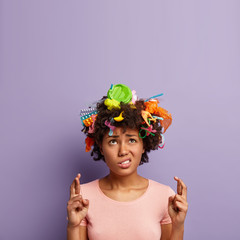 Photo of praying black woman crosses fingers, believes in solving environmental problems, looks sadly upwards, dressed in casual t shirt, has plastic trash in hair, isolated over lilac background