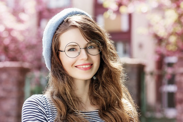 Outdoor close up portrait of young beautiful happy smiling lady wearing round transparent glasses, blue beret. Model posing in street. Spring fashion concept. Copy, empty space for text
