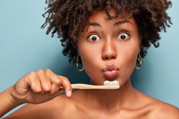 Cropped shot of surprised dark skinned woman with curly hair, keeps toothbrush near folded lips,...