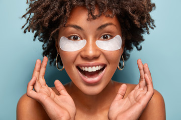 Headshot of ovejoyed beautiful woman reduces under eye puffiness, dark circles, applies moisturizing patches, cares of eye skin beauty, smiles broadly, wants to touch soft face, isolated on blue