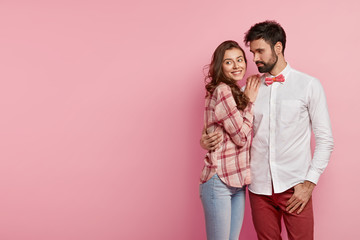 Horizontal shot of happy romatic couple embrace and have pleasant talk, wear stylish elegant clothes, smile happily pose over pink background with free space left for your advertisement or information
