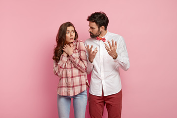 Angry man reproaches girlfriend, gestures and explains something angrily, frustrates puzzled woman keeps hands together, afraids of husbands remarks, sort out relationship, isolated on pink wall