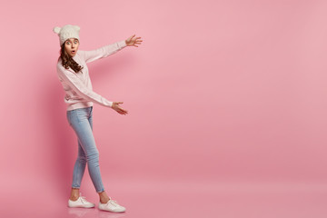 Omg, it is so big. Impressed surprised woman shows big size with hands, overwhelmed to see huge object, wears fashionable headgear, jumper and jeans, gestures over pink background. Free space.