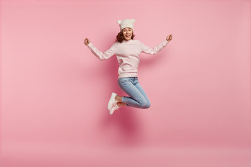 Full length body shot of joyful pleased young female model jumps happily in air against pink background, wears warm hat with ears, sweatshirt, jeans and sneakers, feels energetic and optimistic