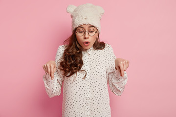 Photo of shocked European lady looks with wonder, points down, says wow, sees surprising item on floor, wears round spectacles, warm hat and shirt, isolated over pink background. Omg concept.