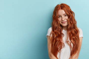 Healthy hair and perfect freckled skin concept. Satisfied teenage girl with red wavy loose hair, gentle smile, wears white t shirt, isolated over blue wall, has natural beauty, poses for making photo