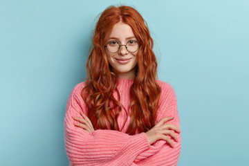 Satisfied ginger charming woman with curly hair, wears transparent glasses, keeps arms folded over chest, has pleasant talk with contemporary, wears oversized clothes, isolated over blue background - obrazy, fototapety, plakaty
