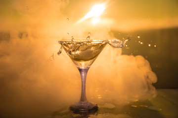 Martini cocktail glass in hand splashing on dark toned smoky background or colorful cocktail in glass with splashes and olives. Party club entertainment. Mixed light. Selective focus