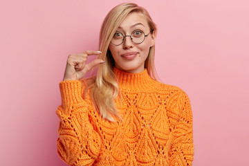Photo attractive light haired curious woman shows little gesture, demonstrates tiny object, tells put bit more efforts to achieve desirable results, looks through spectacles, wears winter jumper