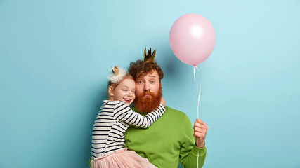 I love you, dad! Bearded redhead father plays with cute little girl, holds balloon, recieves hug from female child, dressed in festive clothes, celebrate Childrens Day together. Family relationships