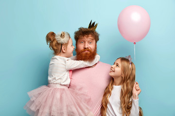Busy father organizes holiday on International childrens day for two daughters, have home party, wear crowns and festive clothes. Little adorable girl holds air balloon, looks at dad and sister - obrazy, fototapety, plakaty