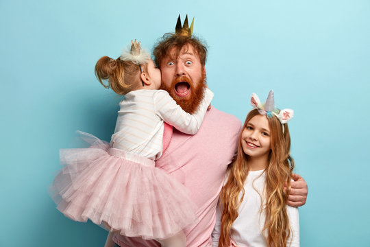 Mad father hears secret from daughter, spends free time with children, embraces and shares love, wear carnival costumes for party, isolated over blue wall. Kids and parents concept. Festive event