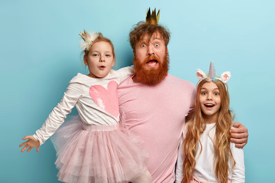 Foxy bearded father stands between two little daughters, embraces and has surprised look, celebrate International Children Day. Impressed dad and two girls with unicorn horn and crowns on heads