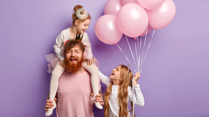 Puzzled tired busy father entertains daughters, feels fatigue of playing different games on party, enjoy birthday celebration stands against purple background. Funny sisters look happily at each other