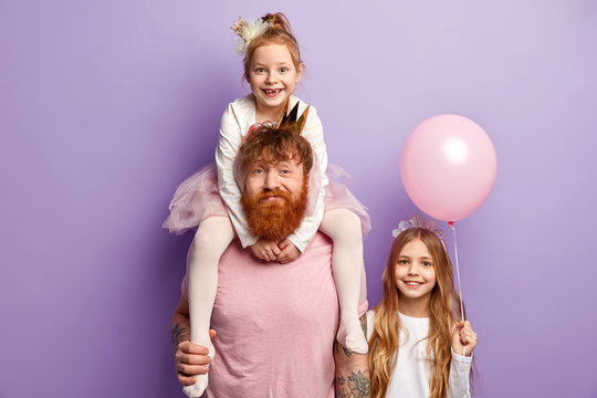 Positive small child on fathers shoulders, celebrates birthday together with dad and sister. Three family members pose against purple background, express different emotions. Children Day concept