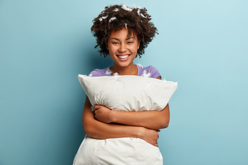 Photo of happy dark skinned woman cuddles white pillow, smiles broadly after awakening in morning,...