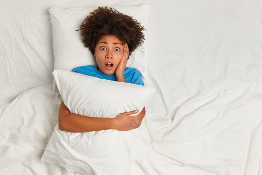 Oh no, I forgot to wake up! View from above of frightened curly Afro American woman embraces pillow, lies in bed on white bedclothes, afraids of dreadful dream, has scared face, stares at camera