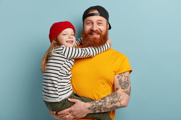 Isolated shot of happy father with ginger beard, yellow t shirt, carries small daughter, recieves...